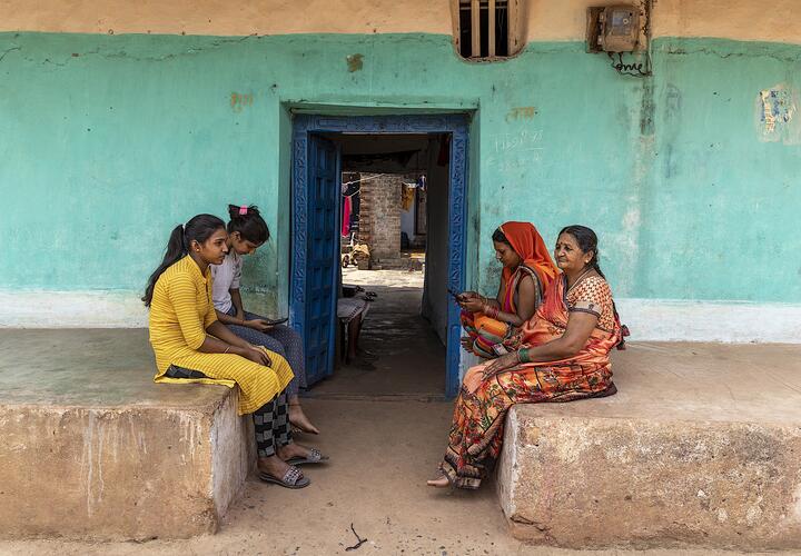 Four Indian women sitting outside, two are using mobile phones. 